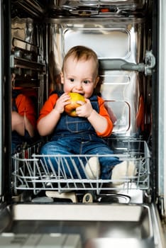 Little baby sitting in empty dishwasher and eating apple