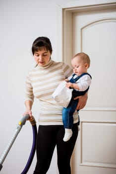 Cleaning up the room - mother with vacuum cleaner holding her baby on hands