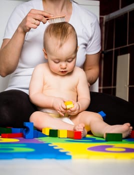 Mother brushing hair of her cute baby while she is playing with toys in bathroom