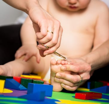 Mother trimming toenails on feet of her baby - toys around