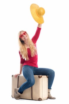 a woman sits on a suitcase and waves with her summer hat