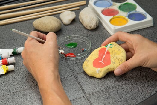 Handmade - stone painting. production decoration. Female hand are drawing red heart on the stone. Horizontally.