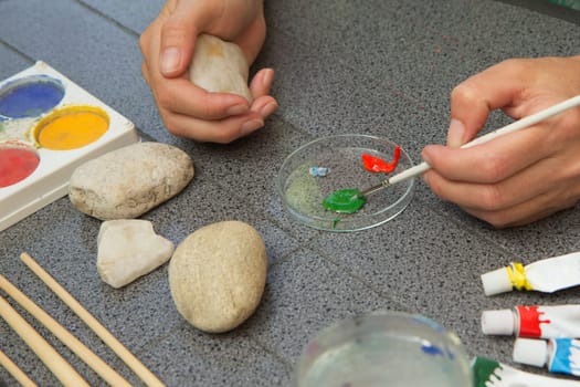 Handmade - stone painting. production decoration. Female hands are dunking brush into the paint. The right hand is holding a clean stone.