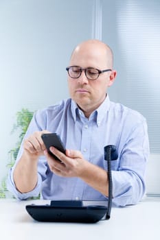 upset upright bald clerk with sunglasses dialing a number on his mobile phone while putting the fixed phone receiver in his pocket
