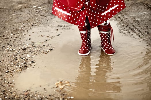 Detail of rainboots of a baby girl dressed with dotted raincoat in the puddles