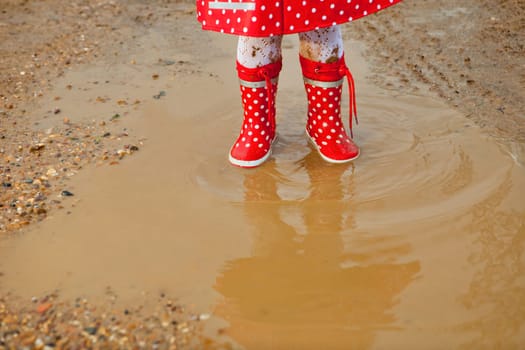 Detail of rainboots of a baby girl dressed with dotted raincoat in the puddles