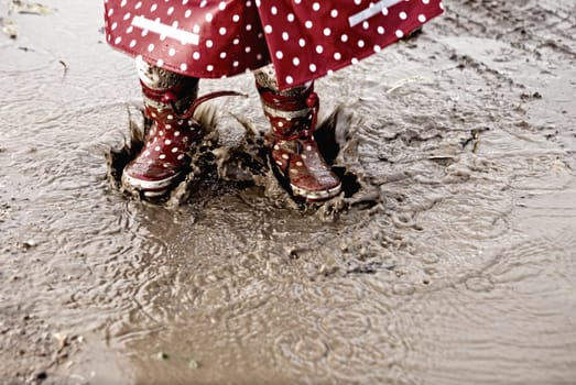 Detail of rainboots of a baby girl dressed with dotted raincoat in the puddles