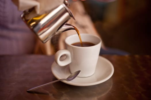 Serving tea from a metal teapont in a white ceramic cup on restaurant terrace table. Selective focus