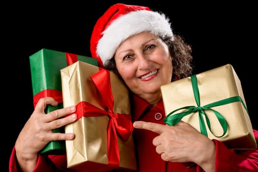 Female senior hugging three wrapped Christmas presents, while pointing her left index finger at two of them. Golden and green wrapping. Santa Claus hat, red coat and white teeth. Isolated on black..