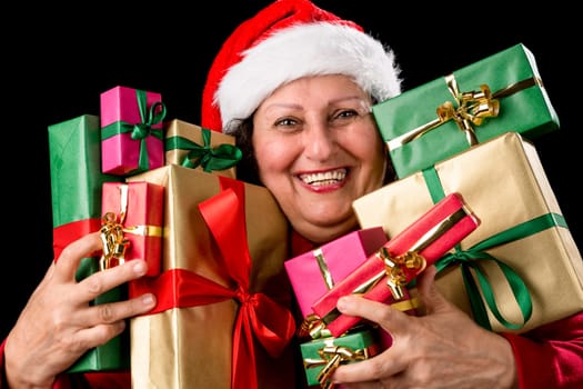 Honorable lady brimming over with joy is pressing ten wrapped Christmas presents to her chest. Santa Claus hat and coat. Smiling teeth. Isolated on black. December theme. Red, green and golden gifts..