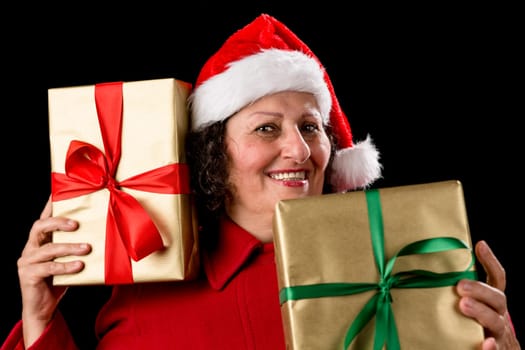 Perky senior woman is holding two gifts, one to her right cheek, the other in front of her chest. Both wrapped in plain golden paper with bows. Red Santa Claus cap. Isolated on black. December theme..