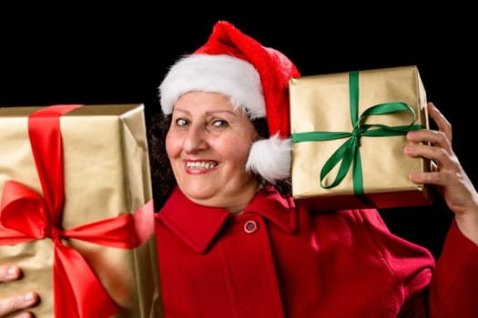 Cheerful mature female with Santa Claus cap and red coat. Holding a gift to her left cheek and another at an arms length. Both Christmas presents wrapped in gold. All isolated on black background..