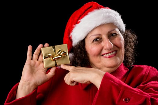 Smiling gentle woman with red coat and Santa Claus cap isolated on black background. Pointing her left index finger at a wrapped golden Christmas present held up by her right hand. Gift-giving theme..