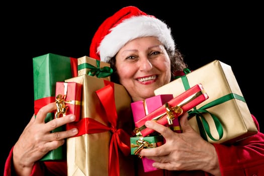 Smiling aged lady with sparkling eyes is embracing eight unicolored wrapped Christmas presents. Santa Claus cap and red coat. White teeth. Isolated on black background. Gifts in gold, red and green..