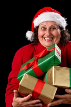 Delighted female pensioner with Santa Claus cap and red coat is carrying three Christmas gifts close to her chest. White teeth. Presents wrapped in plain gold and green. Isolated on black background..