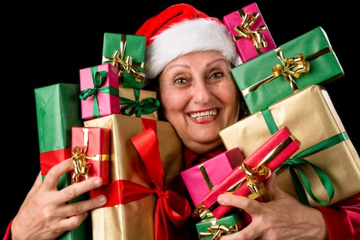Joyful senior lady with Santa Claus cap is embracing a dozen wrapped Christmas presents. All packed in unicolored gold, red and green. Isolated on black background. December gift-giving theme..