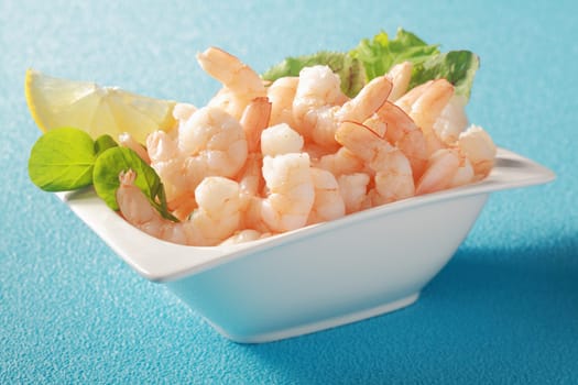 Grilled shelled pink prawns served in a white dish with fresh leafy green herbs as a gourmet appetizer to a dinner, on a blue table