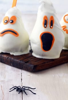 Scary edible Halloween party decorations with fresh pears covered in white chocolate and marzipan to resemble ghosts with spooky expressions on a rustic white table with a spider