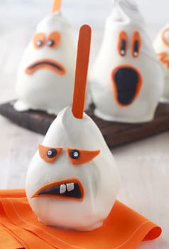 Grotesque Halloween ghost edible decoration with a sneering toothy expression made from a pear coated in white chocolate or marzipan with orange sucker stick and eyes