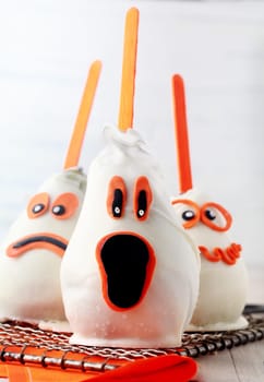 Edible homemade ghost Halloween dessert or party favors made from fresh pears coated in white chocolate and marzipan and decorated with a variety of scary expressions in orange with sucker sticks