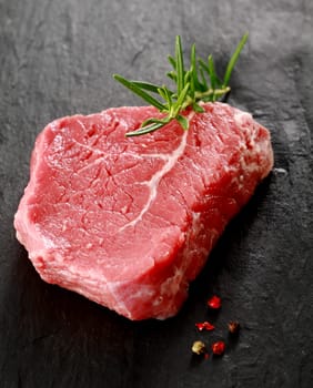 Healthy portion of lean uncooked beef steak garnished with a sprig of fresh rosemary lying on a dark surface with whole peppercorns for seasoning