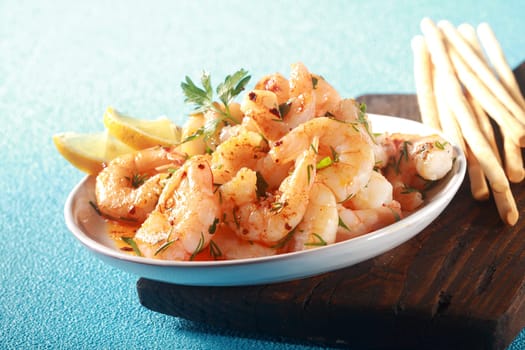 Healthy gourmet appetizer of grilled pink prawns or shrimp seasoned with olive oil and chopped fresh herbs and served with bread sticks on a blue textured table, low angle view