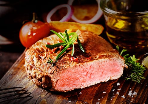 Delicious tender grilled rare beef steak on a chopping board in a rustic kitchen surrounded by fresh ingredients and olive oil and seasoned with rosemary and salt