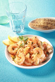Bowl of shelled grilled pink prawns drizzled with olive oil and fresh chopped herbs and served with lemon and bread, high angle view on a blue table