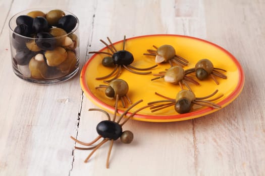 Homemade creative Halloween spider snacks or appetizers formed from green and black olives with Italian spaghetti pasta legs in a realistic shape crawling over a yellow plate on rustic wooden boards