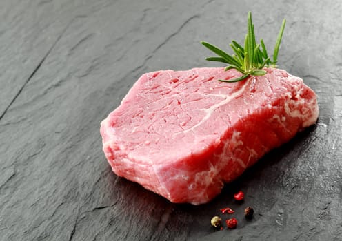 Portion of lean raw beef steak with a sprig of fresh rosemary and peppercorns for seasoning lying on a kitchen counter ready for cooking