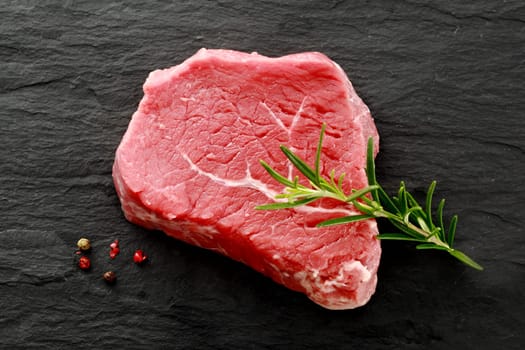 Succulent cut of raw lean rump steak waiting to be cooked with a sprig of fresh rosemary for seasoning and whole peppercorns, view from above on a dark kitchen counter