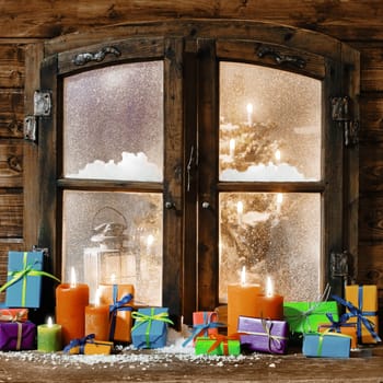 Colorful Christmas gifts and glowing multicolored candles on a windowsill of a rustic timber cabin with a view through the snowy frosted window of a decorated Xmas tree