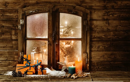 Festive wooden Christmas cabin window with gift-wrapped colorful orange presents, burning candles and decorations in winter snow and a glimpse of a decorated Christmas tree through the frosted window