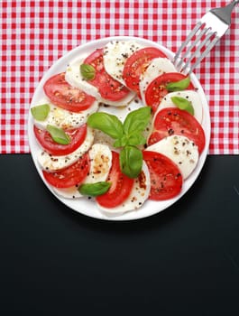 Overhead view of a plate of Italian Caprese salad with alternating slices of tomato and mozzarella cheese seasoned with basil and pepper on a two tone background with red and white check and dark grey