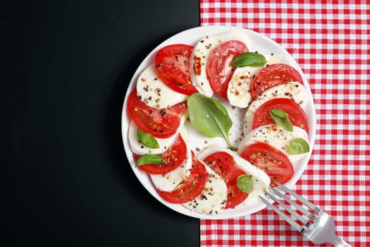 View from above of a plate of Italian Caprese salad with colorful alternating slices of tomato and mozzarella cheese seasoned with basil and pepper on a red and white check and dark grey background