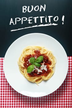 Overhead view of a plate of spaghetti Bolognaise or Bolognese under a Buon Appetito handwritten sign with a rich tomato and ground beef sauce garnished with basil and parmesan cheese