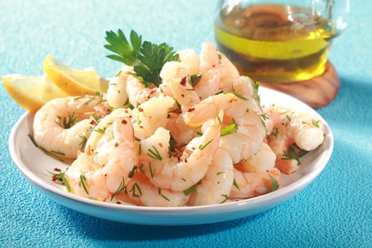 Grilled pink shrimps seasoned with fresh herbs and drizzled with olive oil served in a dish with lemon as a delicious seafood delicacy, on a textured blue table