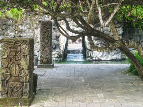 Detail of a pool in Bali with small cave with waterfall and religious symbols.