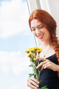 Thoughtful young woman looks out the window in summer with flowers in her hand
