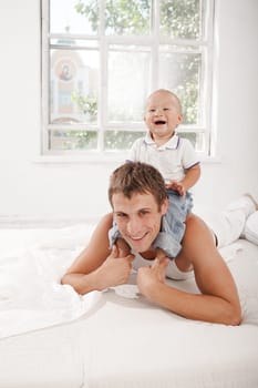 Young smiling father with his nine months old son on the bed at home on white home background. son sitting on his father&#39 s shoulders