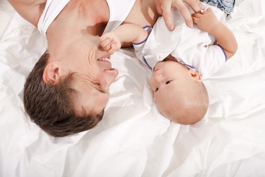 Young smiling father with his nine months old son  lying upside down on the bed at home on white home background
