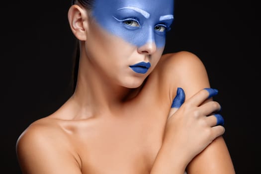 Portrait of a young woman who is posing covered with blue paint in the studio on a black background. girl&#39 s hand around the face and painted blue