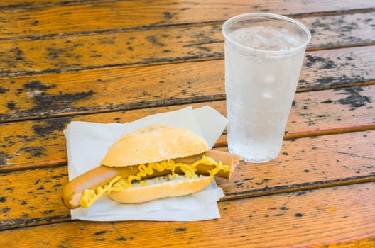 sausage with mustard in bread roll and a cup of mineral water on a brown wooden table on a white napkin.