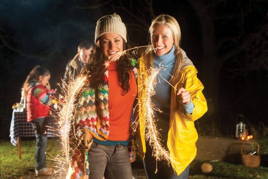 Two women are standing together smiling for the camera with lit sparklers in their hands. They are outside and there are other people in the background behind them.