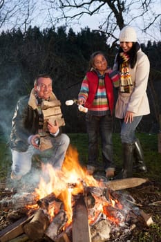 girl and her parents with a bonfire. The father is building up the fire with logs wilst the girl is holding marshmallows ready to roast with her mother.