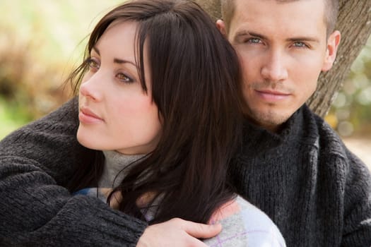 Attractive young couple cuddling out doors. The man is looking at the camera whilst his partner is looking away to the side.