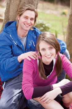 Young couple sitting on a tree trunk outside, smiling at the camera
