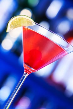 A cosmopolitan, or informally cosmo, is a cocktail made with vodka, triple sec, cranberry juice, and freshly squeezed lime juice or sweetened lime juice. On the photo - cosmopolitan cocktail in front, blurred bottles of alcoholic drinks in the background.