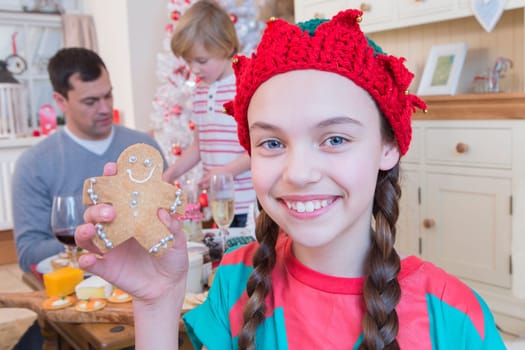 Young Girl dressed as an Elf at Christmas Time with Dad and Brother in the background. She is smiling at the Camera and holding up a decorated Gingerbread Man.