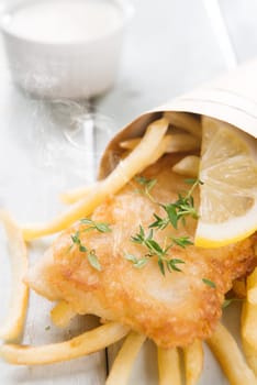 Fish and chips. Fried fish fillet with french fries wrapped by paper cone, on wooden background. Fresh cooked with hot steams.